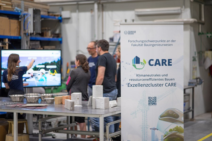 © André Terpe Interessierte Menschen hören sich eine Präsentation einer jungen Wissenschaftlerin vor einem großen Bildschirm an. Auf dem Tisch im Vordergrund liegen ein paar Exponate aus Beton und alternative Bewehrungen.